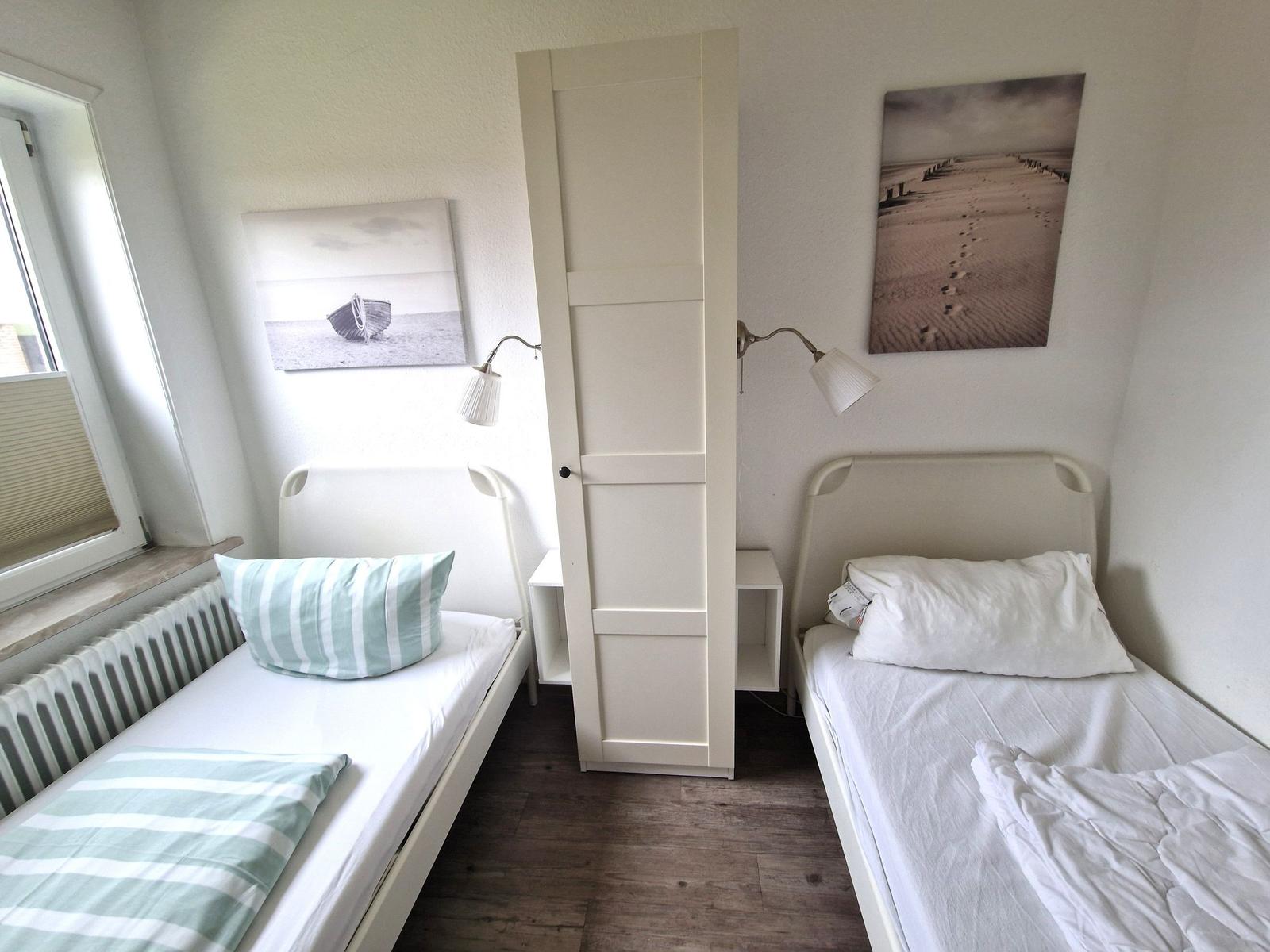 Zwei Einzelbetten, weiße Wände, Wandbilder und Schrank im Raum.