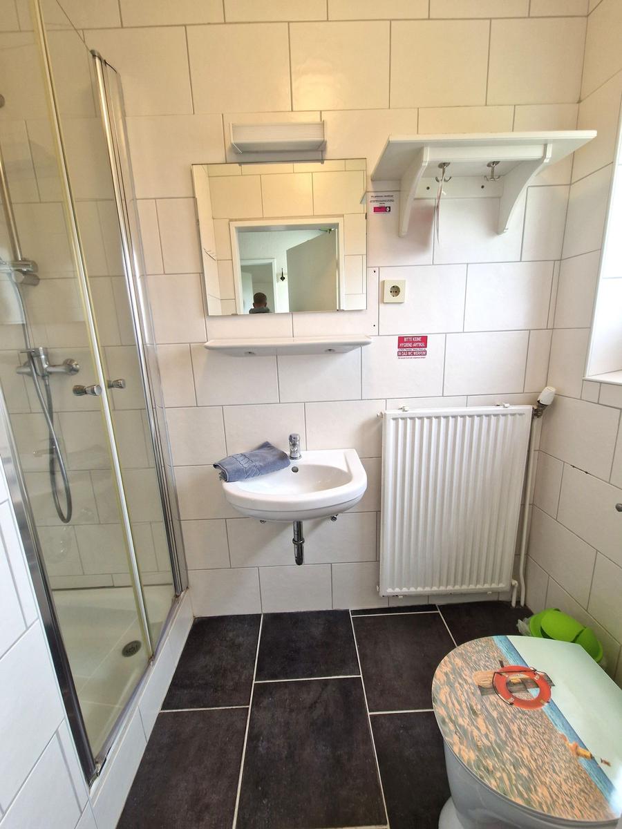 Badezimmer mit Dusche, Waschbecken und Toilette. Weiße Fliesen und dunkle Bodenfliesen.