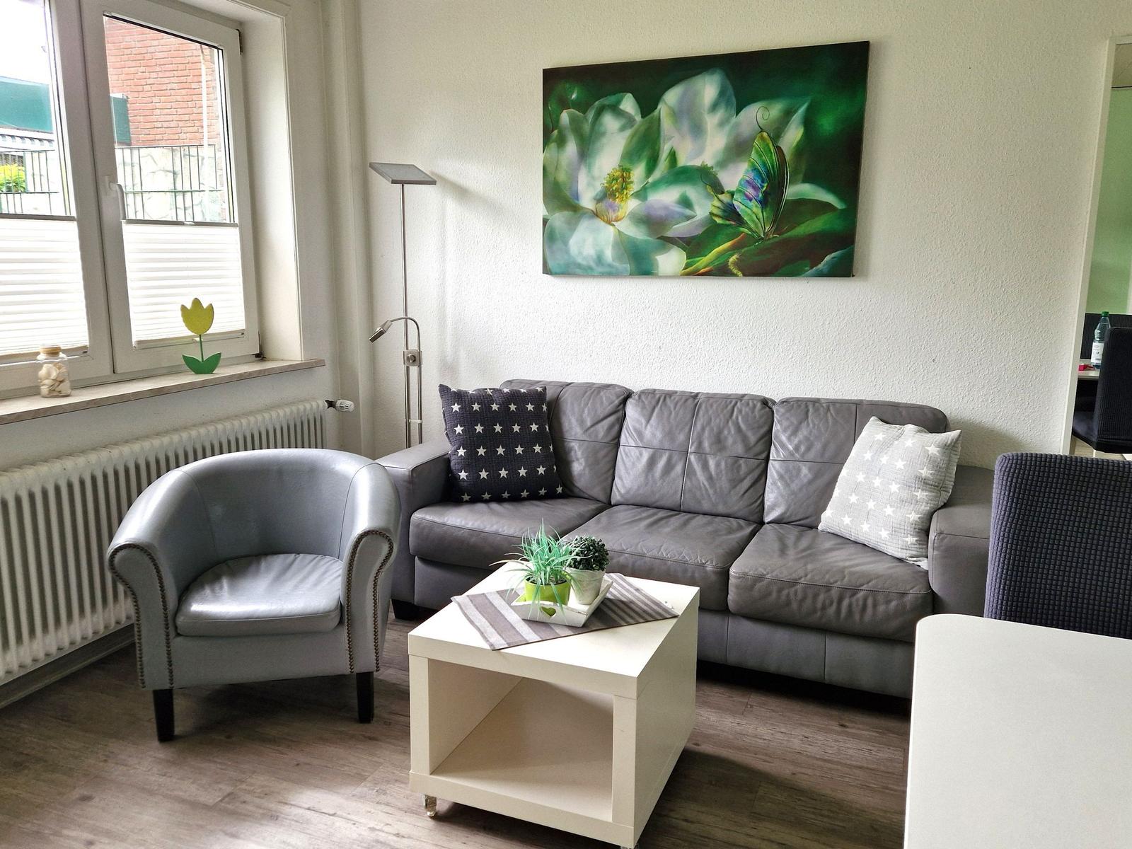 Wohnzimmer mit grauem Sofa, Sessel und Wandbild. Fenster mit Jalousie und Heizkörper.