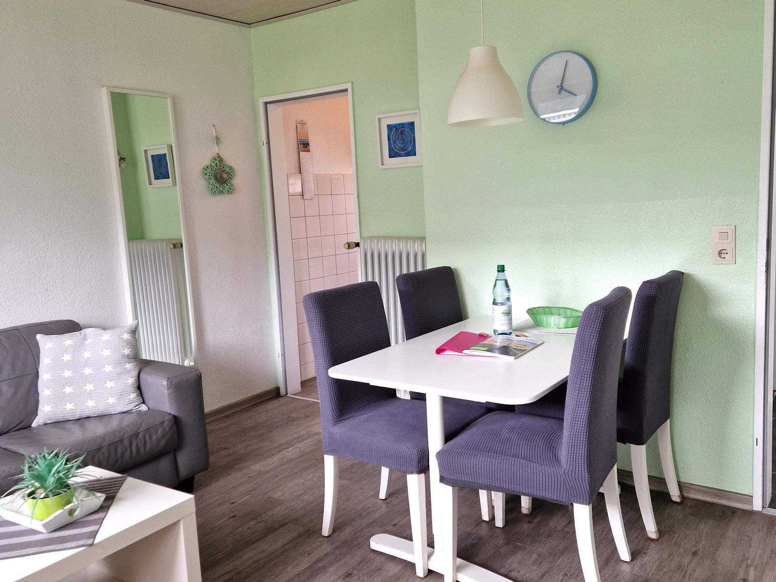 Essbereich mit Tisch und Stühlen, Sofa und Wanduhr. Hintergrund: Tür und Spiegel.