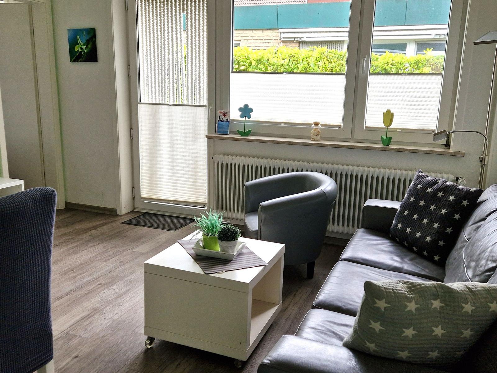 Wohnzimmer mit Sofa, Sessel und Tisch. Fenster mit Jalousie und Blumen dekoriert.