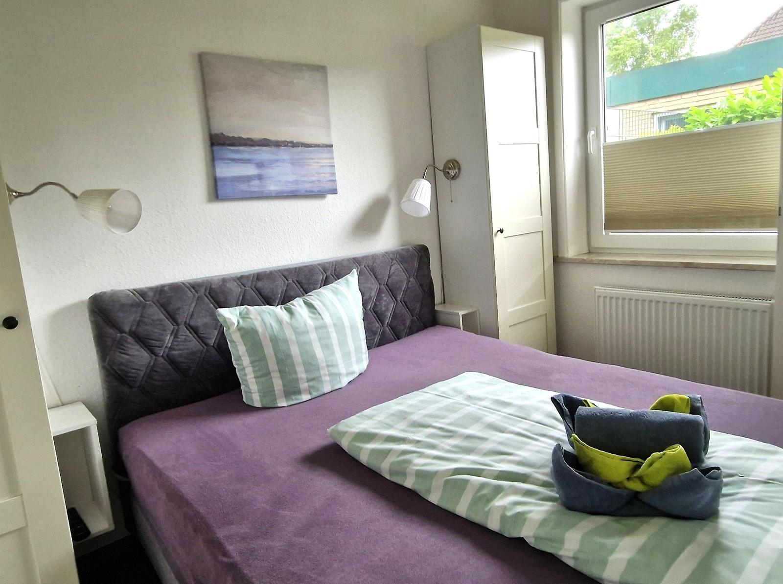 Schlafzimmer mit Bett, Wandbild und Fenster. Auf dem Bett liegen Handtücher.