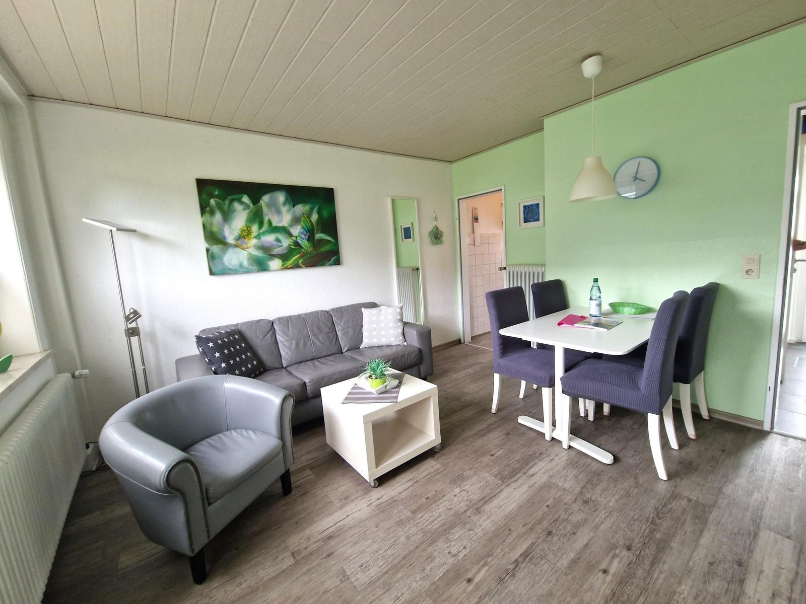 Wohn- und Essbereich mit Sofa, Sessel, Tisch und Stühlen. Grüne Wand, Holzboden und Wandbild.