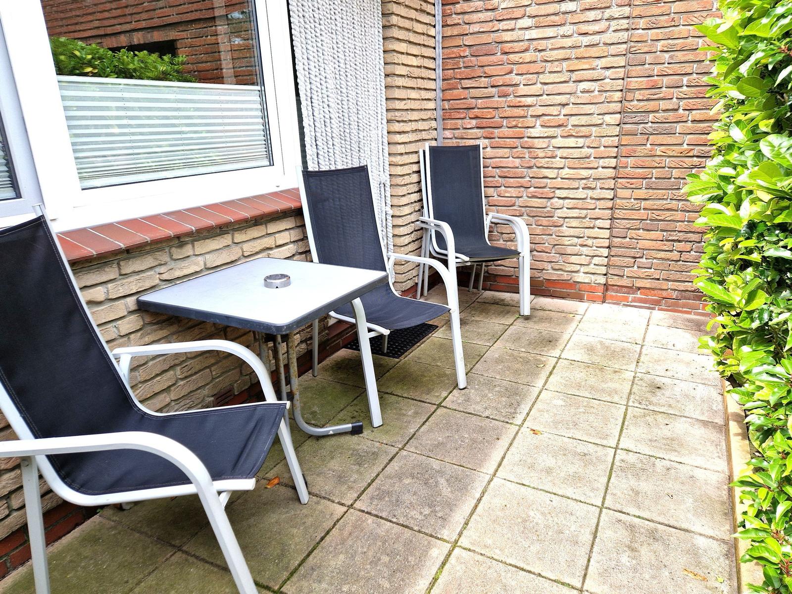 Terrasse mit Tisch und drei Stühlen, Wand aus Ziegelsteinen, Pflanzen und Fenster im Hintergrund.
