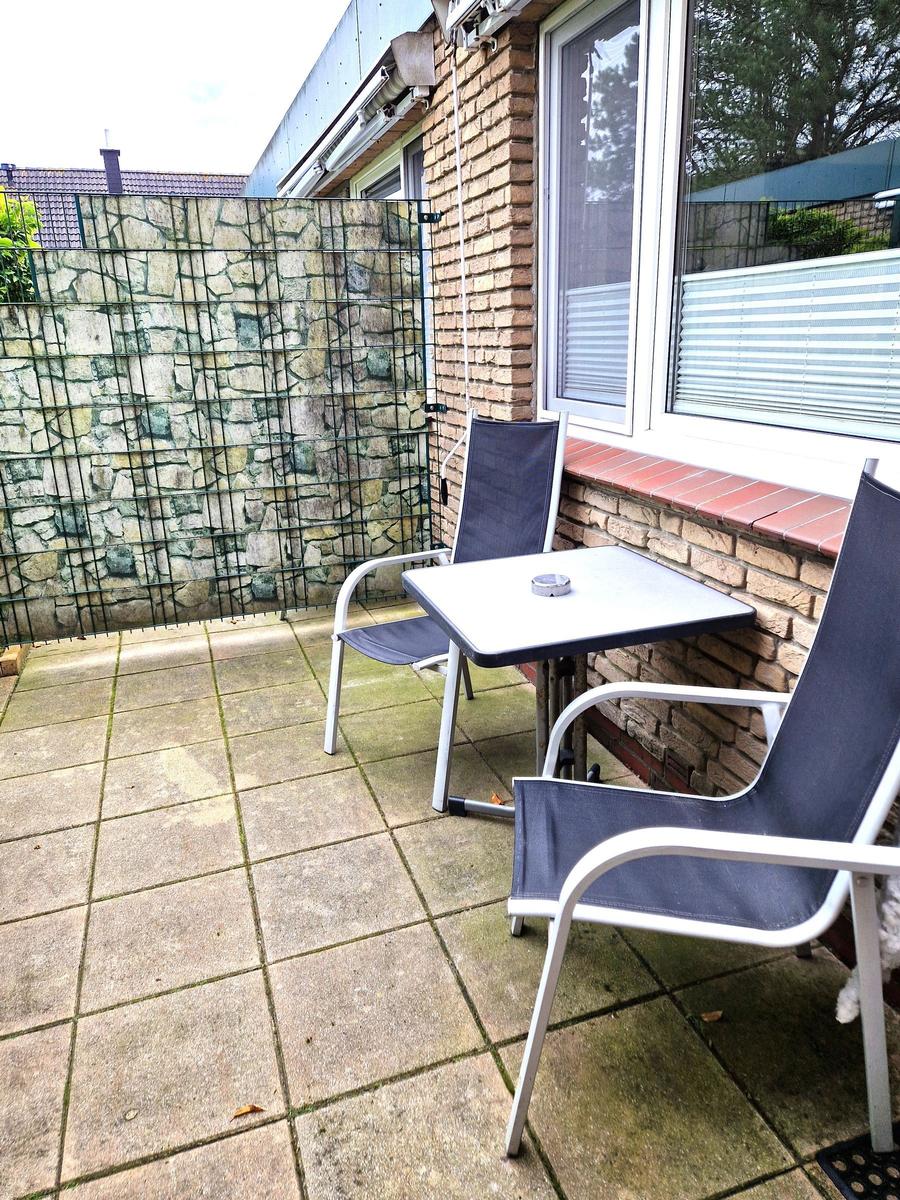 Terrasse mit Tisch und zwei Stühlen, Wand aus Stein und Metallgitter.