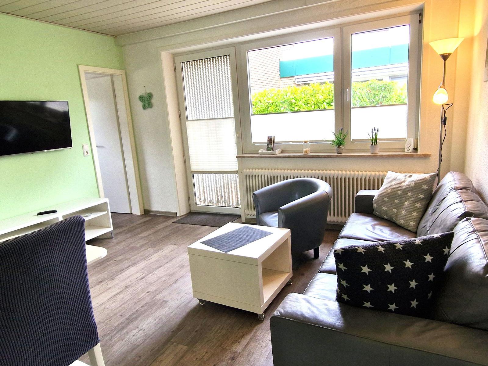 Wohnzimmer mit Sofa, Sessel, Fernseher und Fenster mit Blick auf Grünfläche.