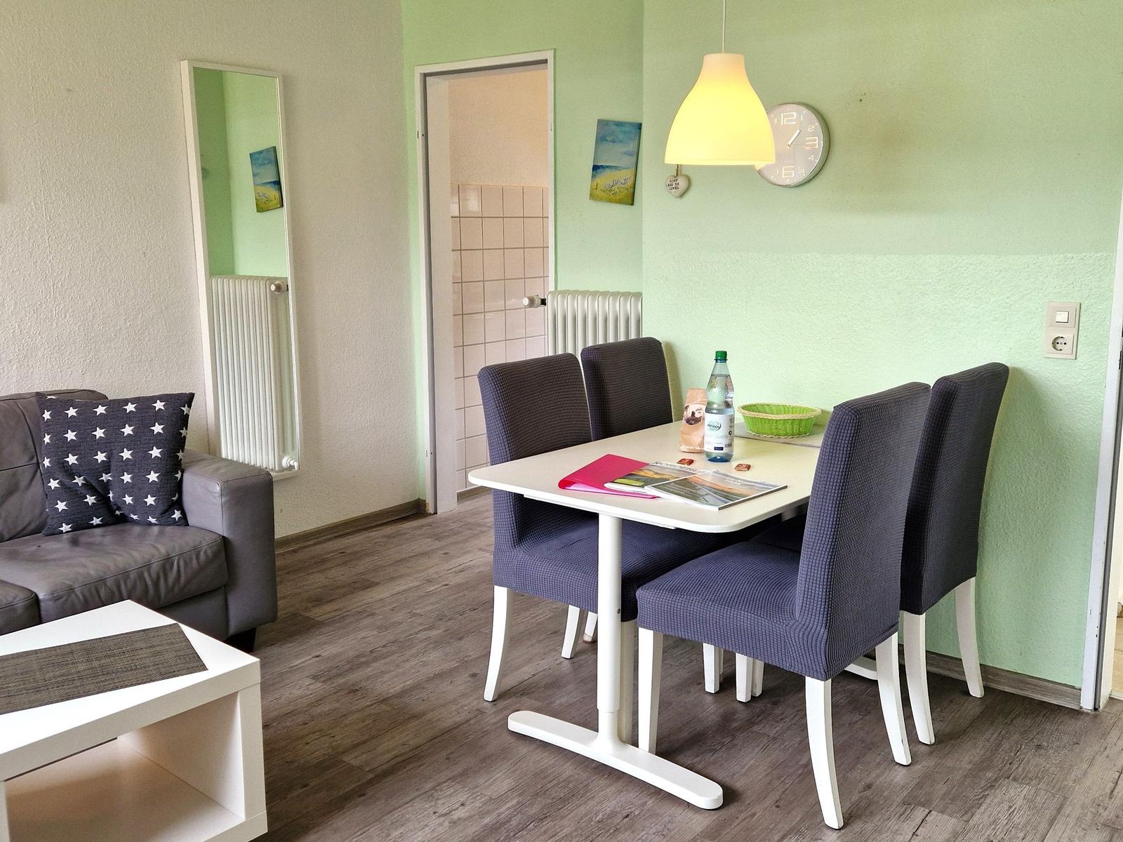 Essbereich mit Tisch und Stühlen, Sofa mit Kissen, Wanduhr und Deckenleuchte.