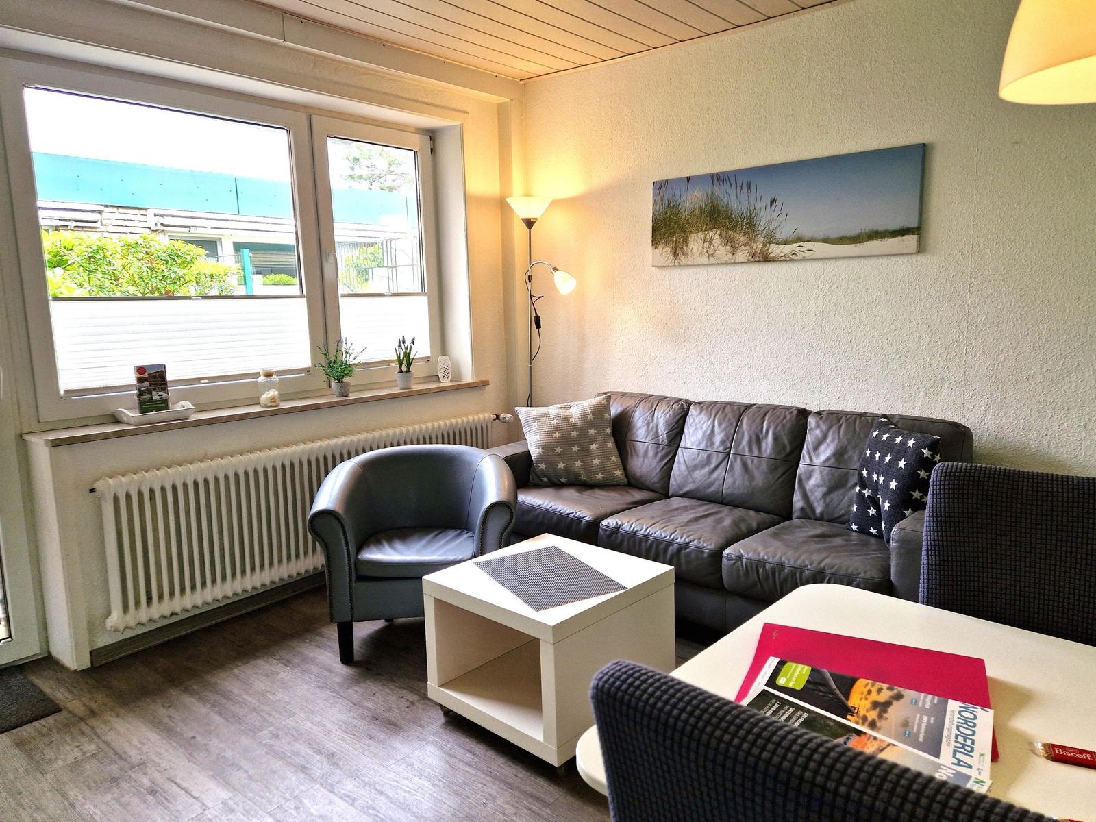 Wohnzimmer mit Sofa, Sessel und Tisch. Fenster mit Blick auf Außenbereich.