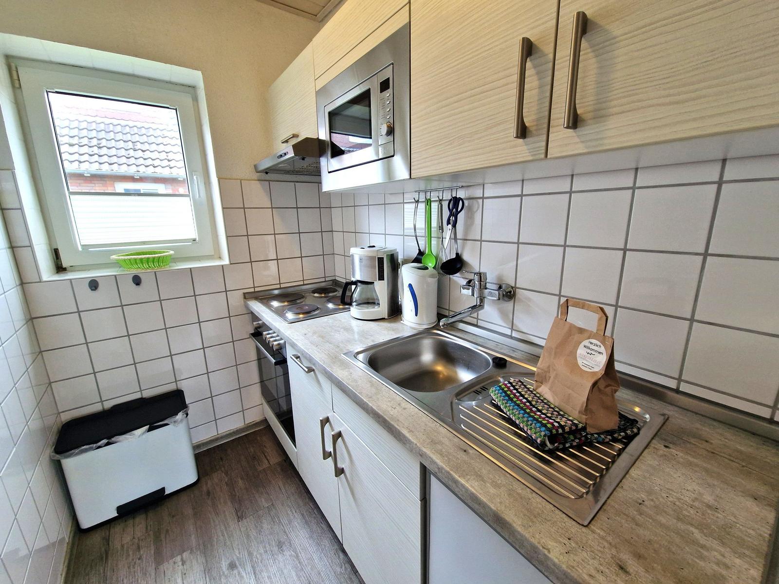 Küche mit Spüle, Herd, Mikrowelle und Fenster. Auf der Arbeitsplatte steht eine Tüte.