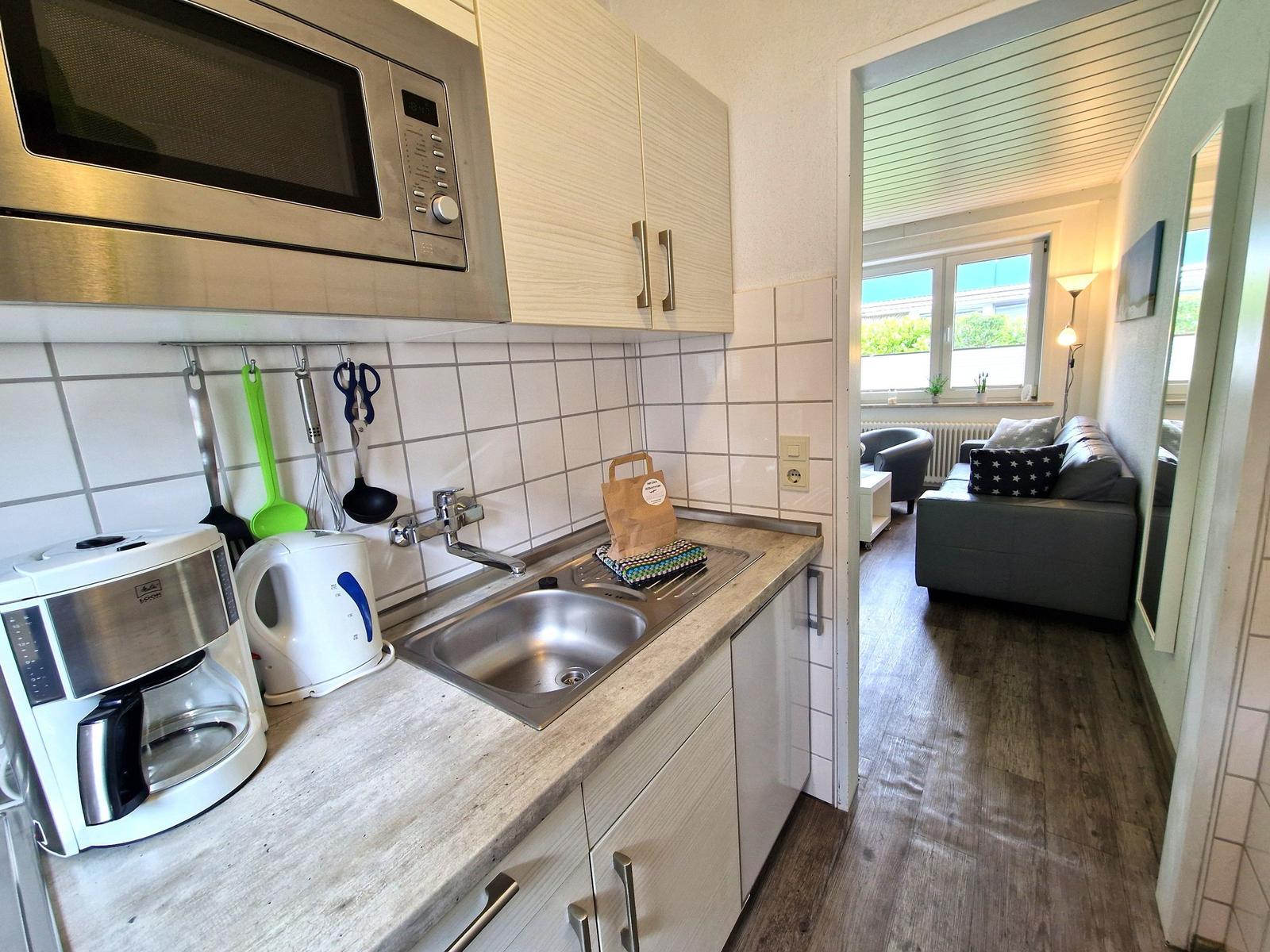 Küche mit Spüle, Mikrowelle und Kaffeemaschine. Durchgang in Wohnzimmer mit Sofa.
