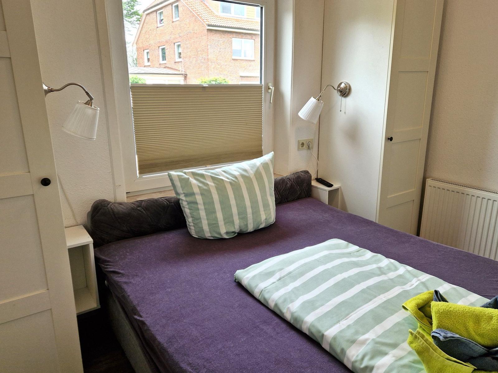 Schlafzimmer mit Bett, Fenster und Wandlampen. Auf dem Bett liegt eine Decke.