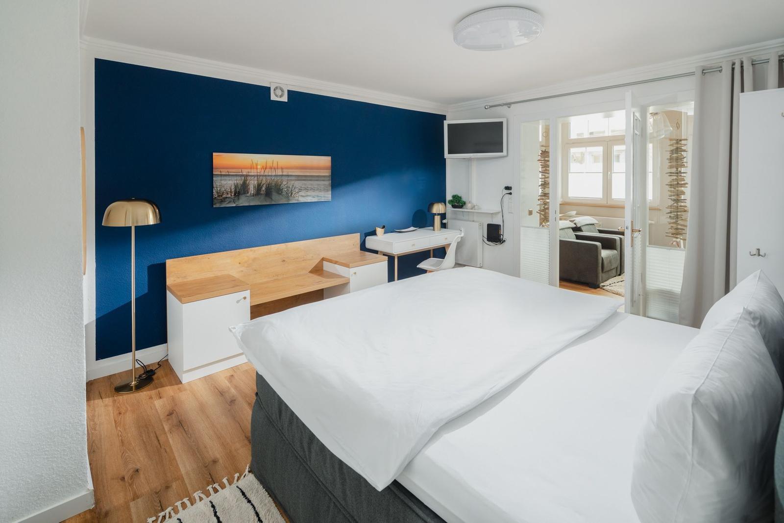 Schlafzimmer mit blauer Wand, Bett, Schreibtisch und TV. Durchgang zu Wohnbereich.