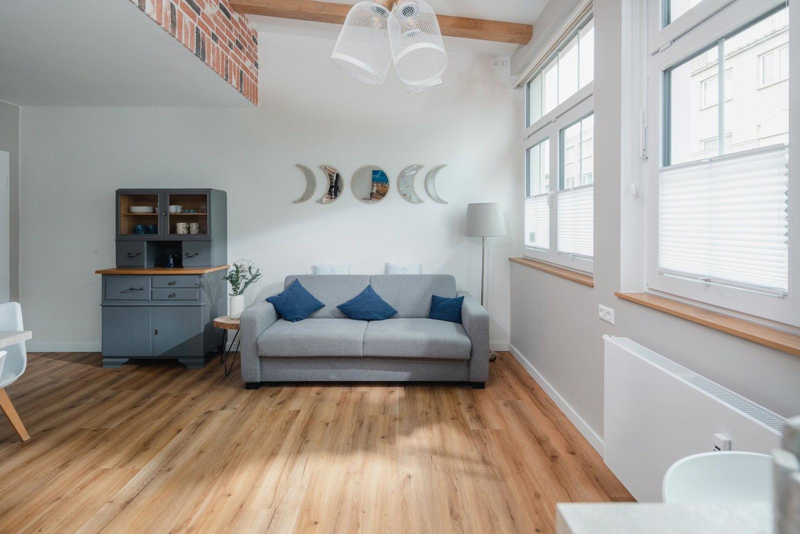Wohnzimmer mit grauem Sofa, Holzboden und Fenstern mit Jalousien.