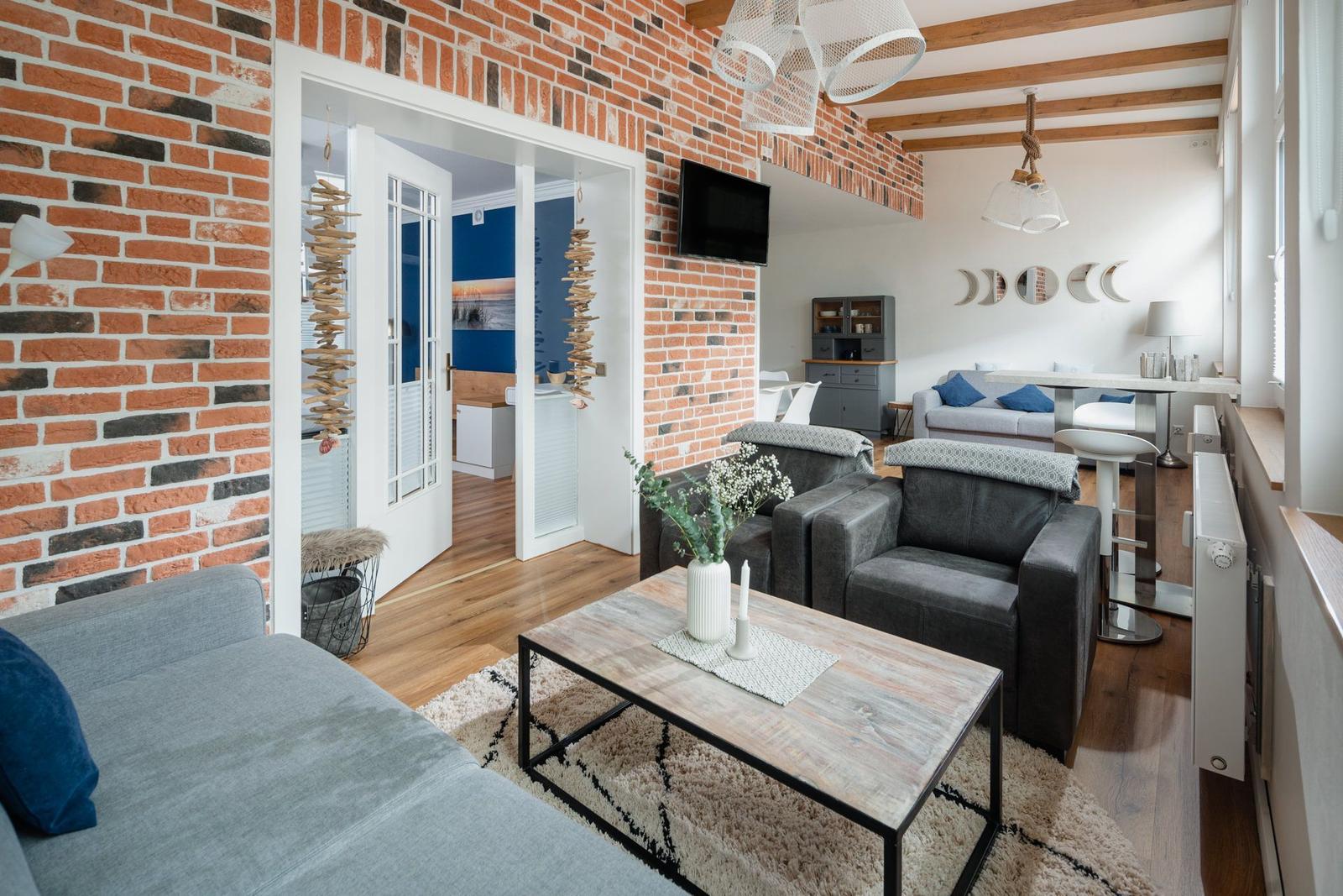 Wohnbereich mit grauen Sesseln, Holzboden und Backsteinwand. Küche und Schlafzimmer sind sichtbar.