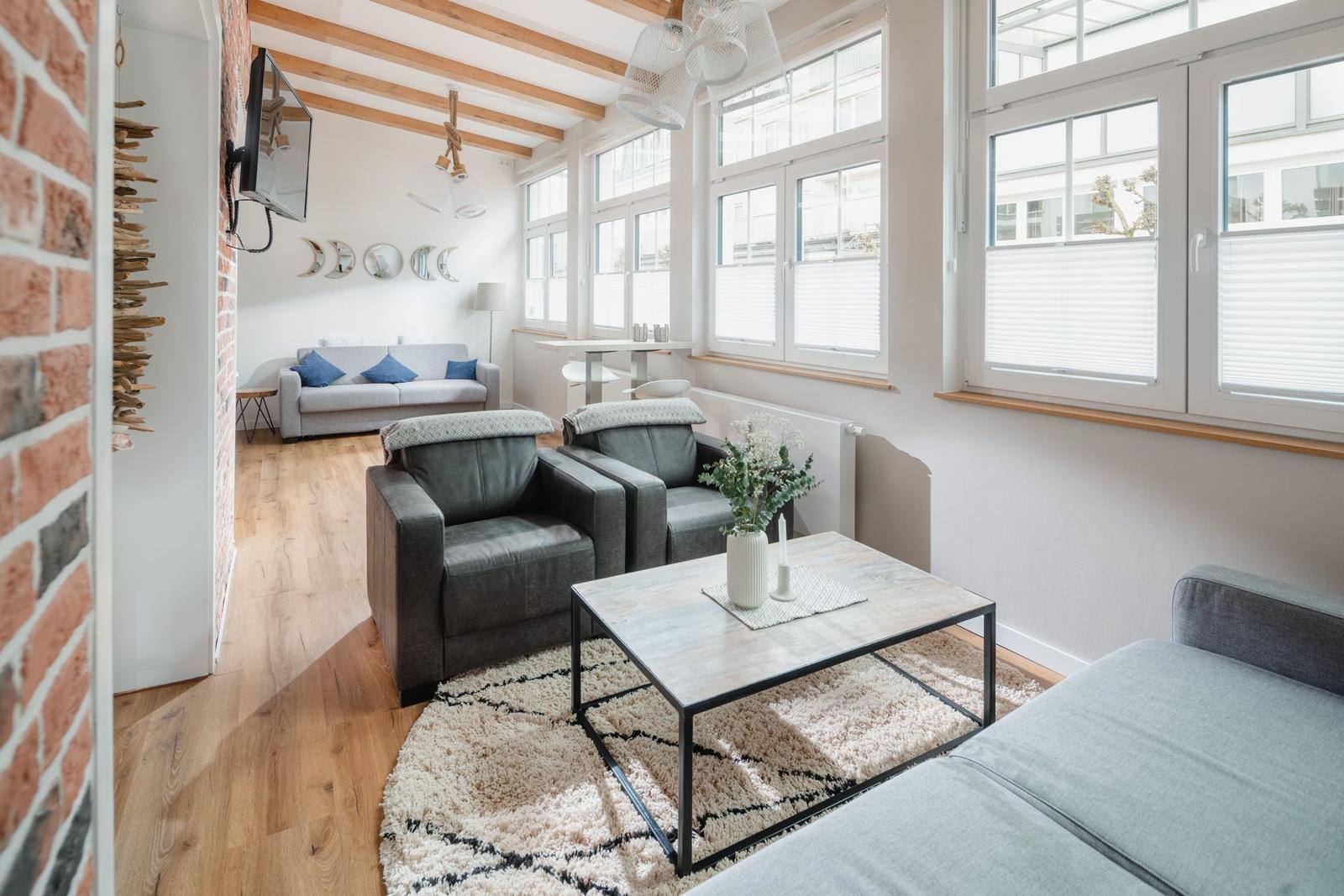 Wohnzimmer mit grauen Sesseln, Couch und Holzboden. Große Fenster und Deckenbalken.