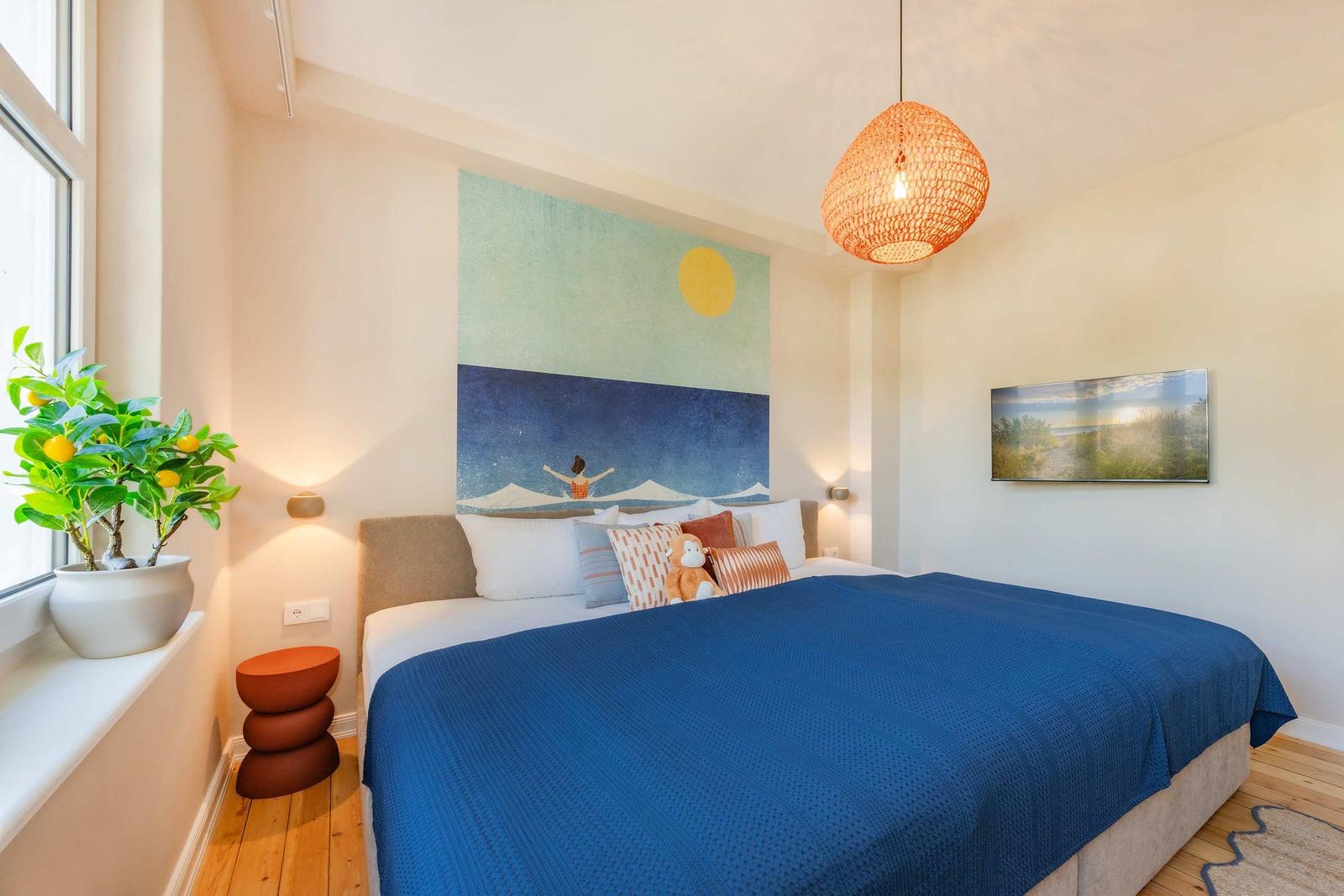 Schlafzimmer mit Bett, Wandbild und Pflanze am Fenster.