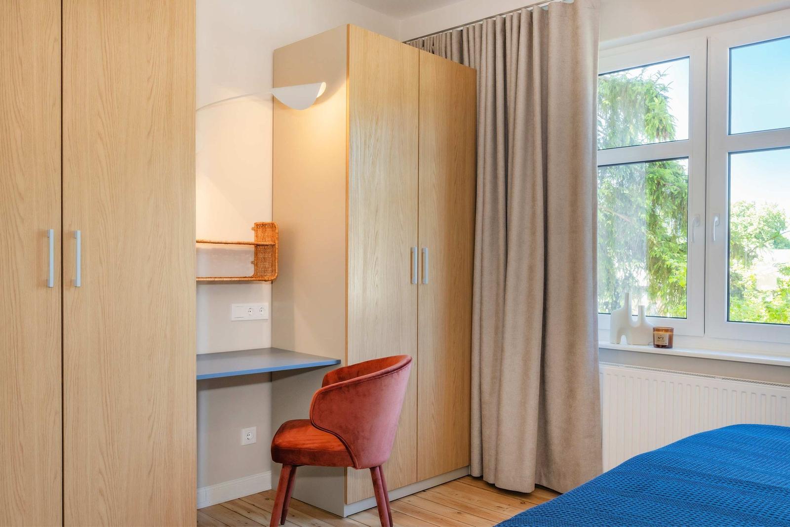 Ein Schlafzimmer mit Schreibtisch, Stuhl und Kleiderschrank. Fenster mit Vorhängen und Blick auf Bäume.