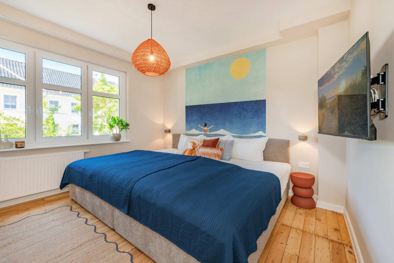 Schlafzimmer mit Bett, Wandbild, TV und Fenster mit Blick auf Außenbereich.