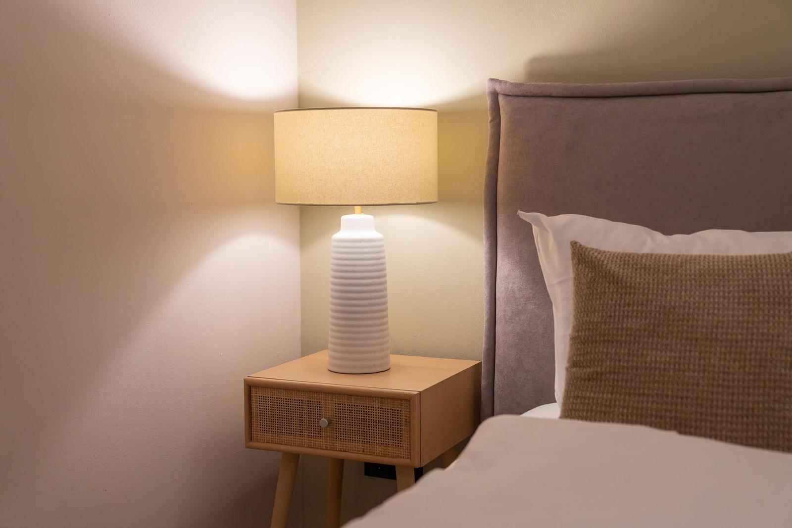 Bettkopfteil, Nachttisch und Lampe sind sichtbar. Die Lampe ist eingeschaltet.