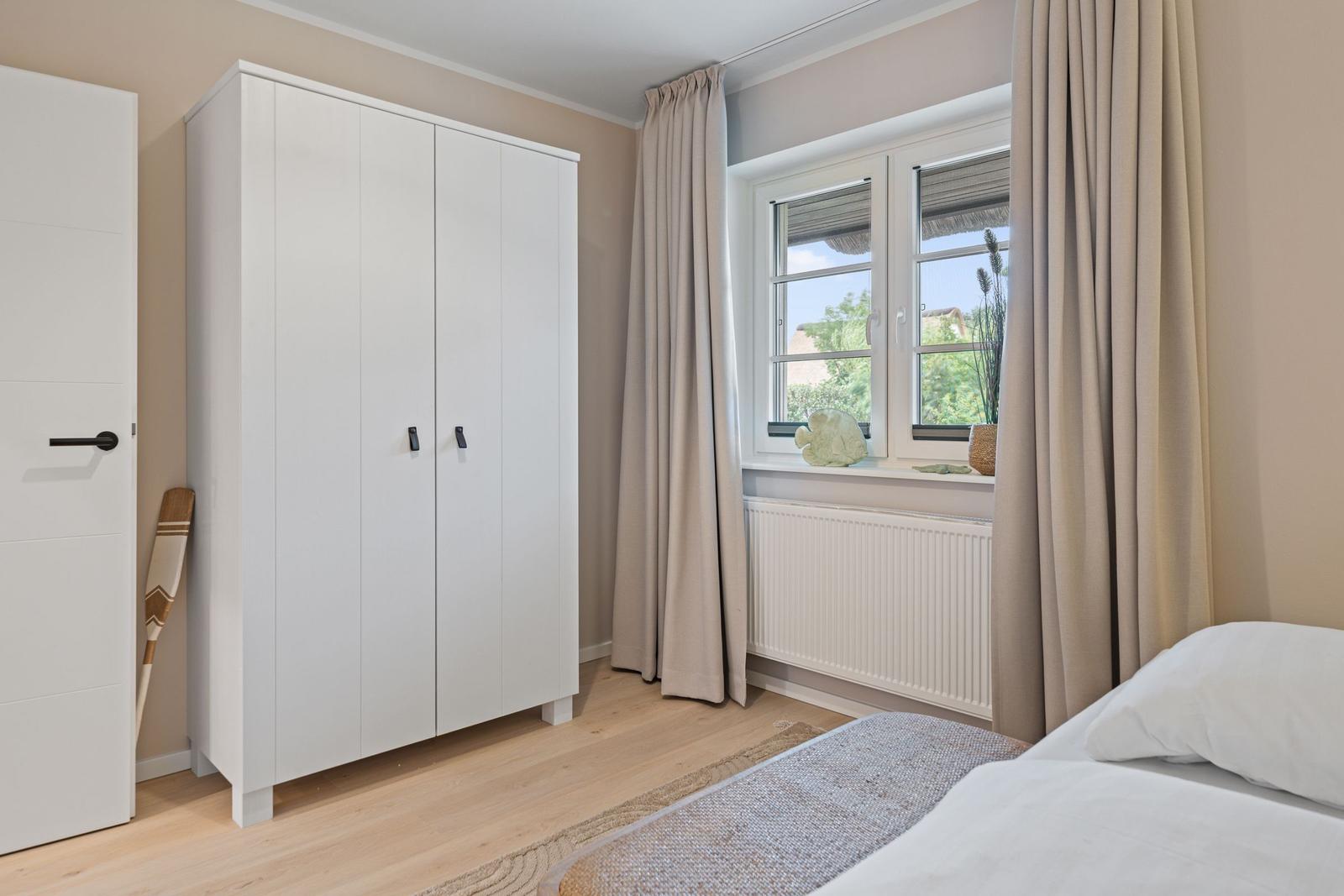 Schlafzimmer mit weißem Schrank, Fenster mit Vorhängen und Bett mit weißer Bettwäsche.