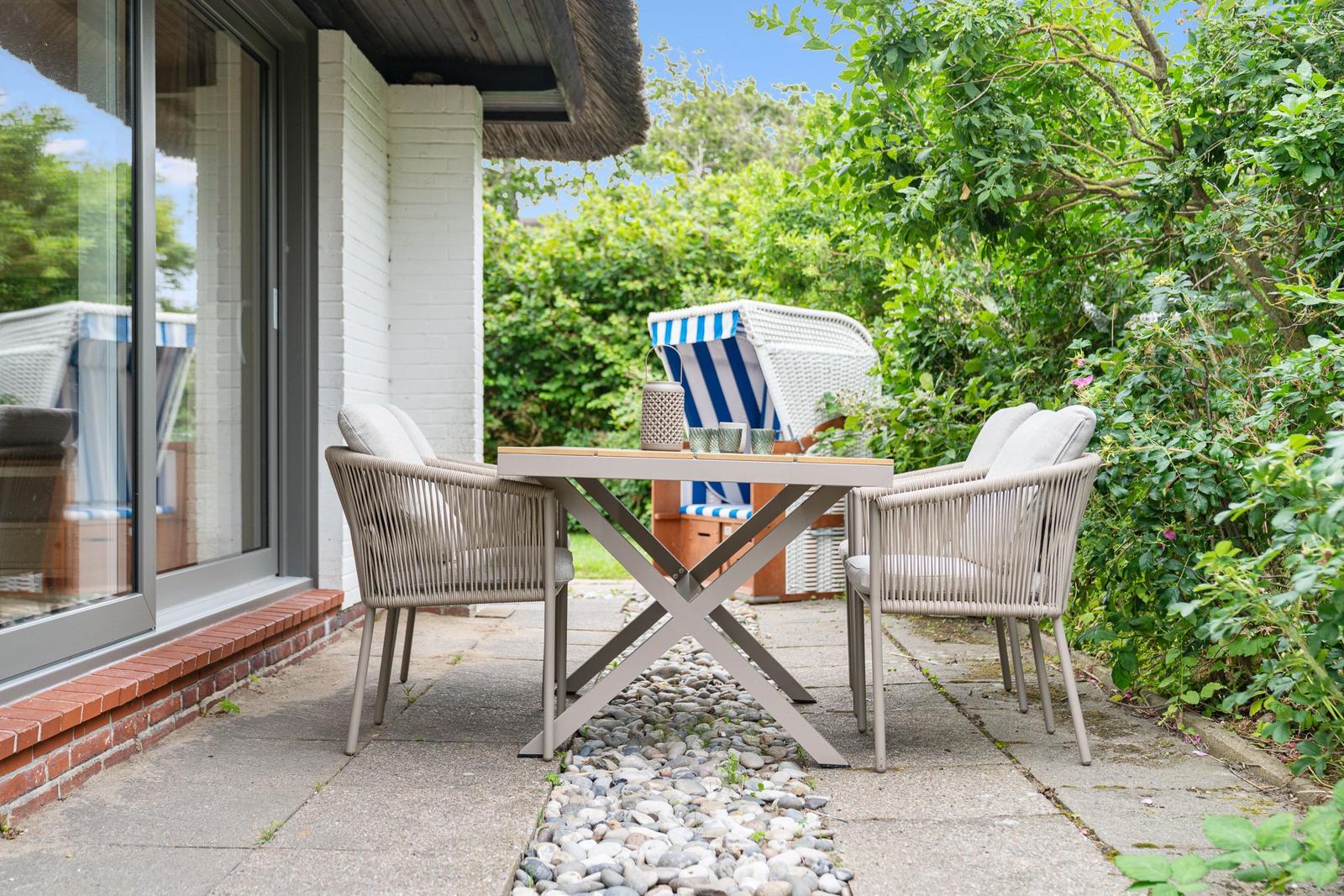 Terrasse mit Tisch und Stühlen, Strandkorb und grüner Umgebung.
