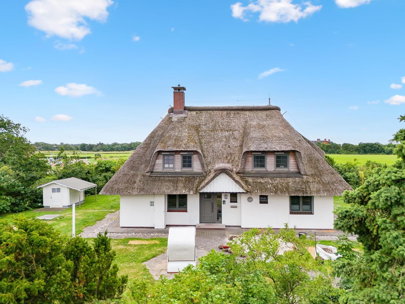 Haus mit Strohdach, weiß verputzt, umgeben von Grünflächen und Bäumen unter blauem Himmel.