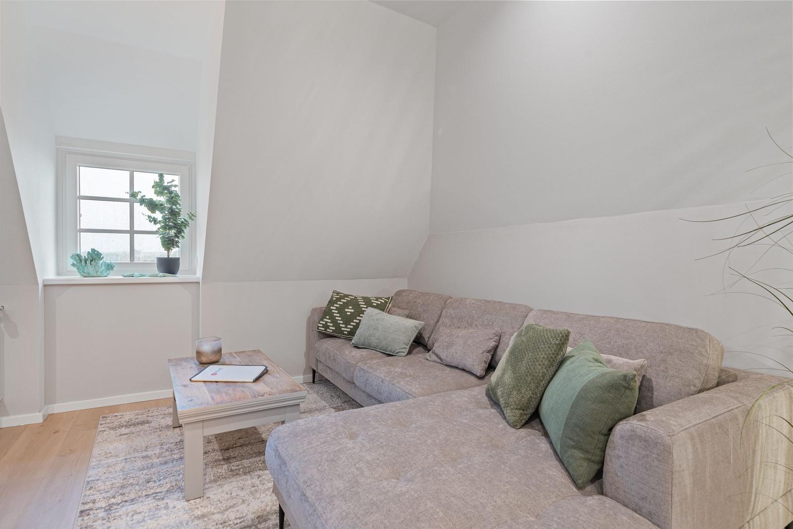 Ein Wohnzimmer mit Sofa, Tisch und Fenster. Grüne Kissen und Pflanzen sind sichtbar.