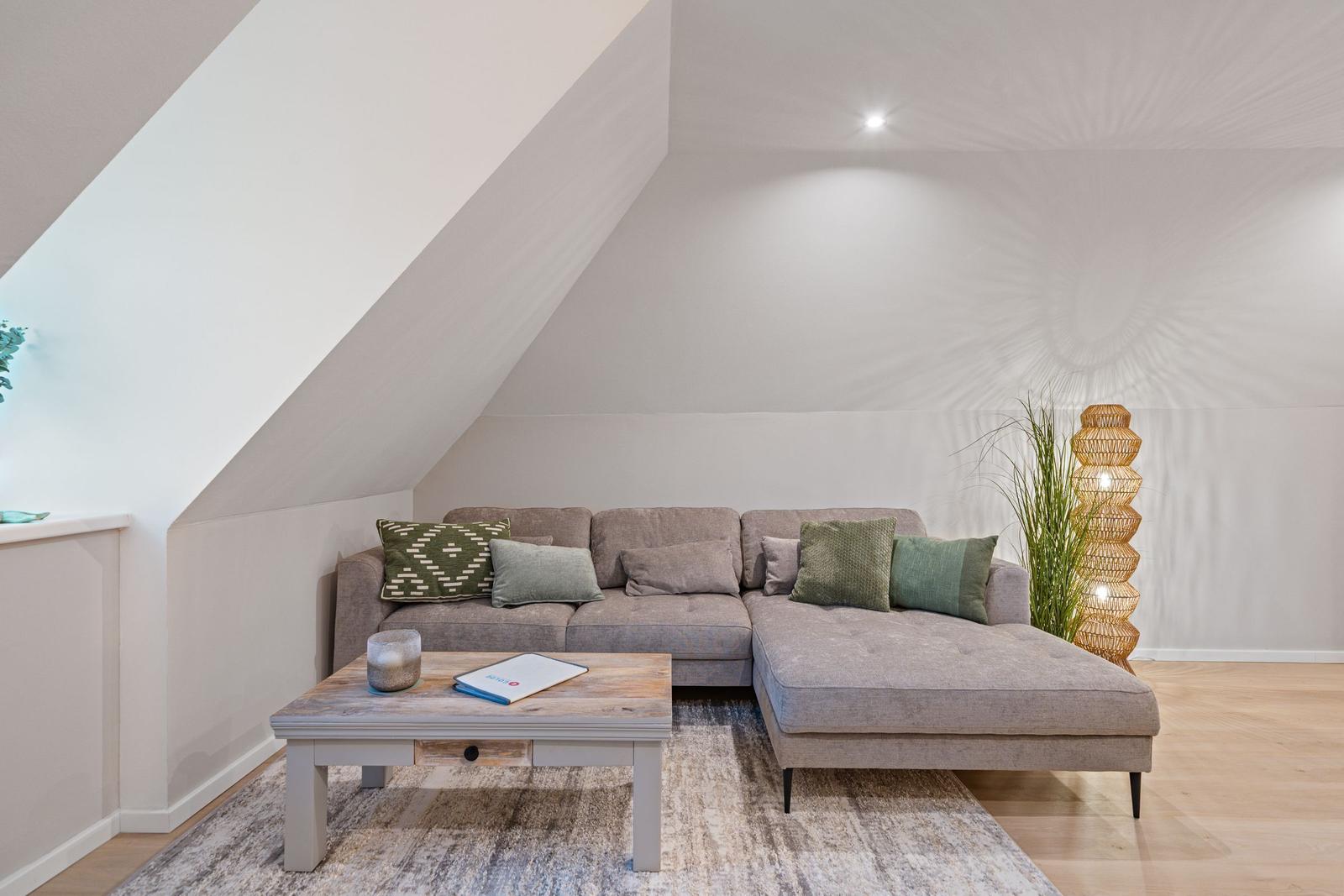 Ein Wohnzimmer mit grauem Sofa, Holztisch und Wandleuchte unter Dachschrägen.