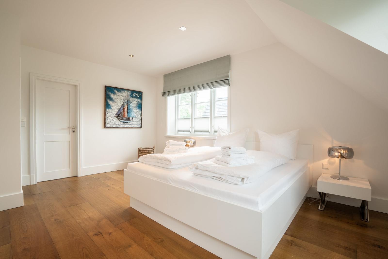 Schlafzimmer mit weißem Bett, Holzboden und Fenster. Wandbild zeigt Segelboot.