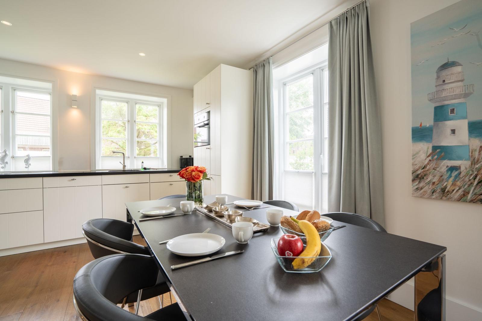 Essbereich mit Tisch, Stühlen und Obst. Küchenbereich mit Fenstern und Wandbild.