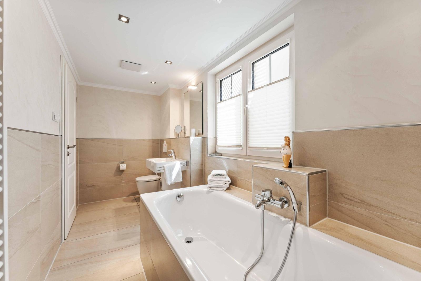 Bad mit Badewanne, Waschbecken und Toilette. Fenster mit Jalousien und Handtuchstapel.