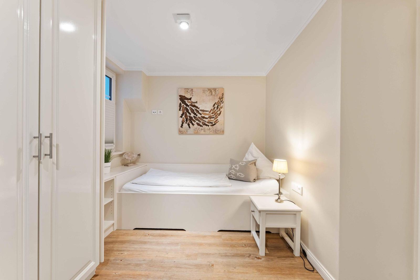 Ein Schlafzimmer mit weißem Bett, Nachttisch und Kleiderschrank. An der Wand hängt ein Bild.