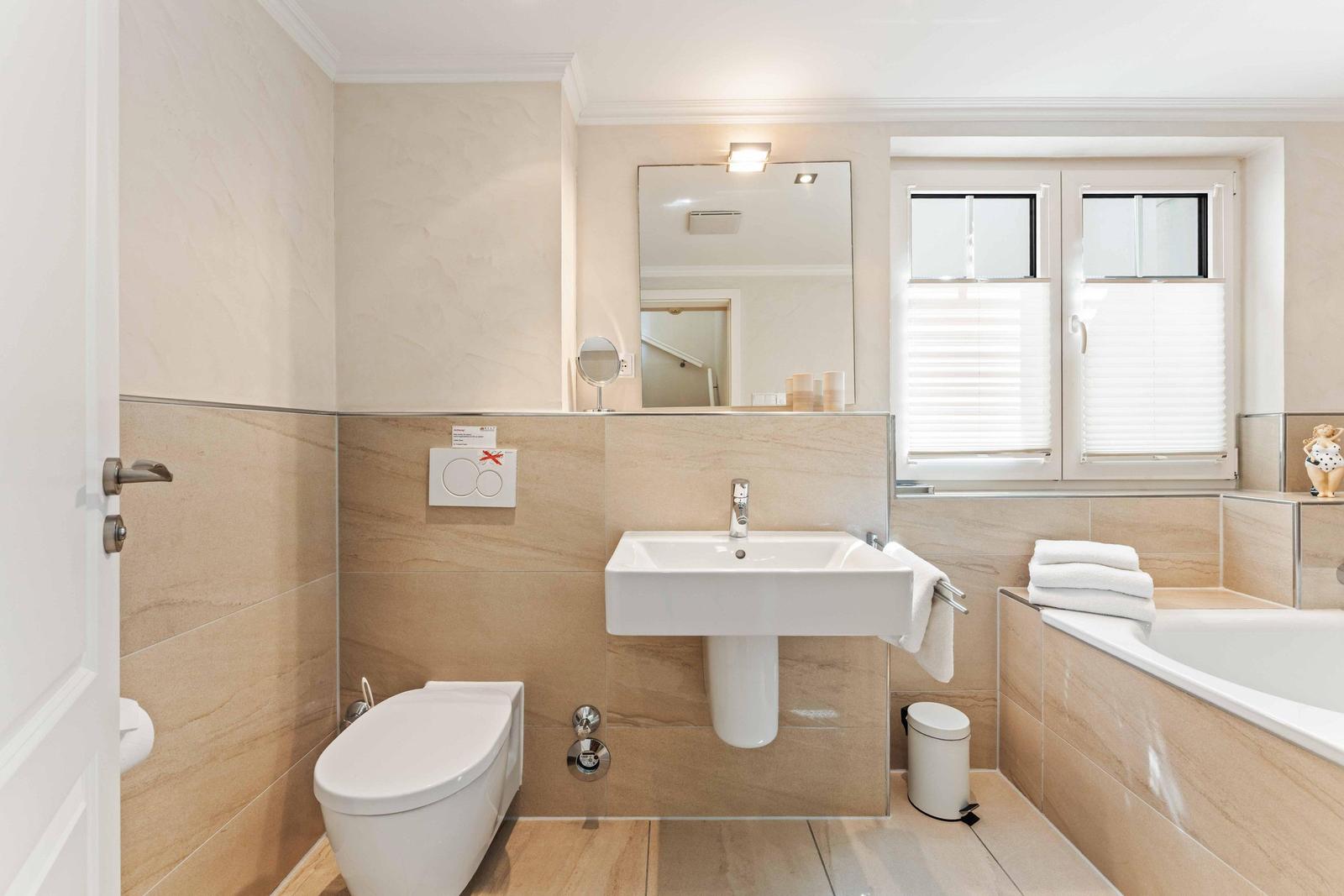 Badezimmer mit Toilette, Waschbecken und Badewanne. Fenster mit Jalousien und Spiegel.