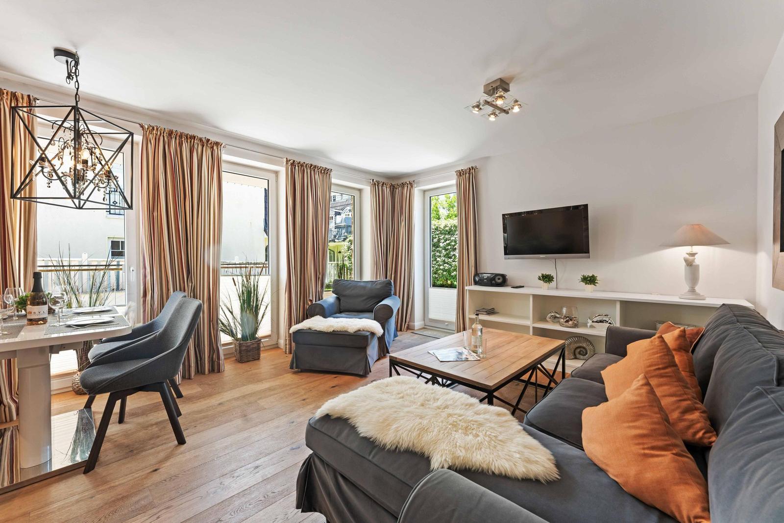 Living and dining area with sofa, coffee table, and TV. Wooden floor and large windows.