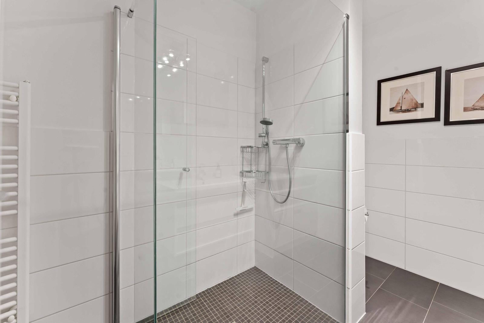 Shower with glass enclosure, white tiles, and handheld shower. Two pictures hang on the wall.
