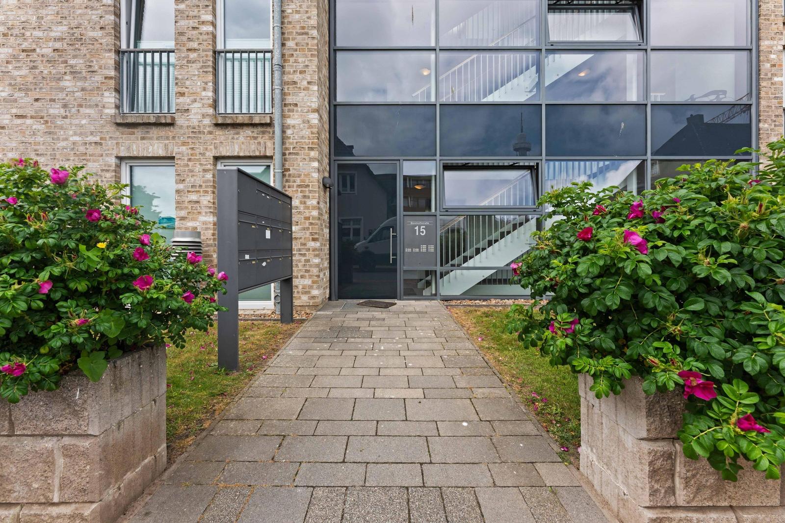 Paved path leads to glass-fronted entrance with balcony. Both sides bordered by rose hedges.
