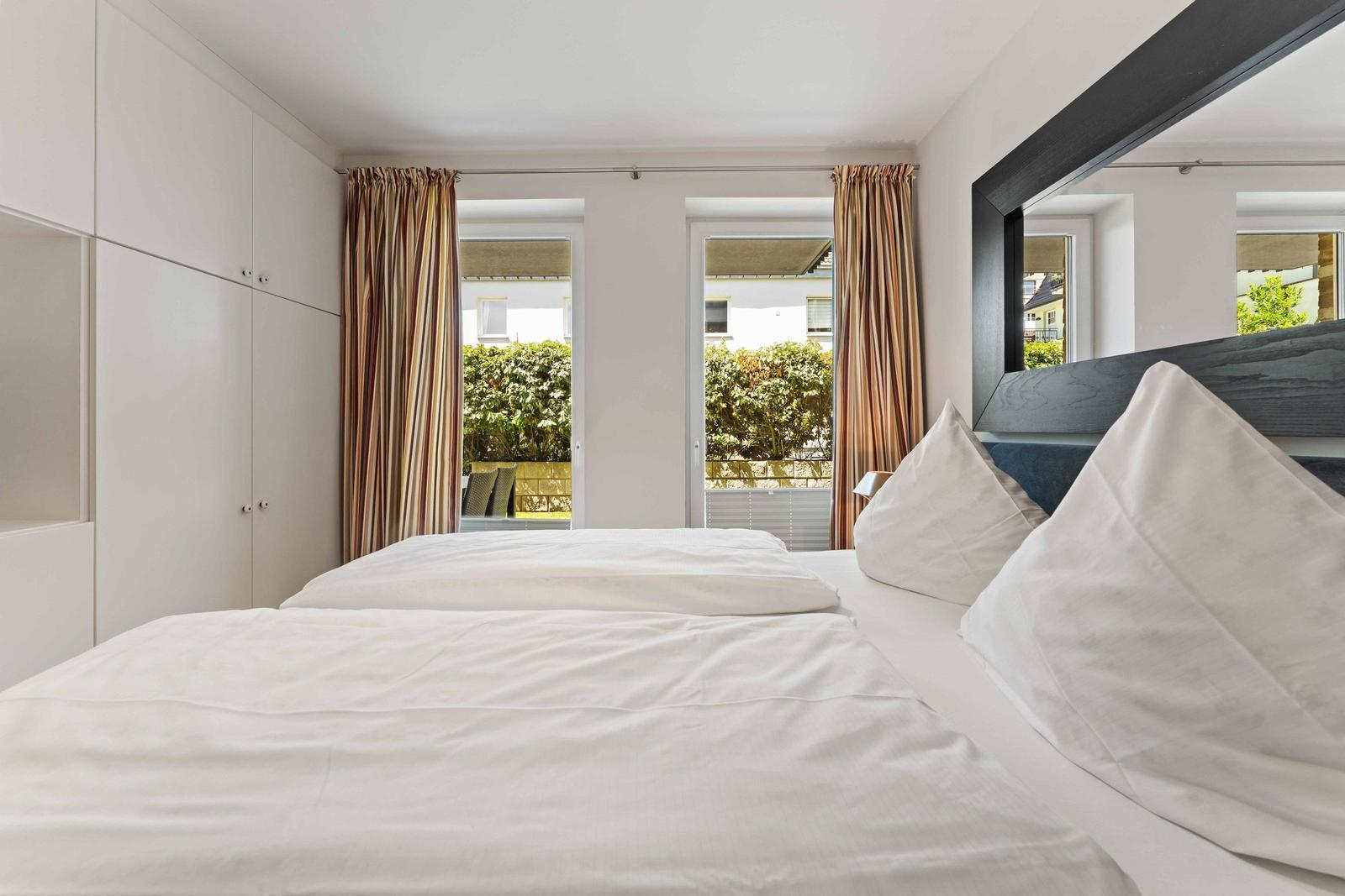 Bedroom with double bed, white linens, and view of balcony with greenery.