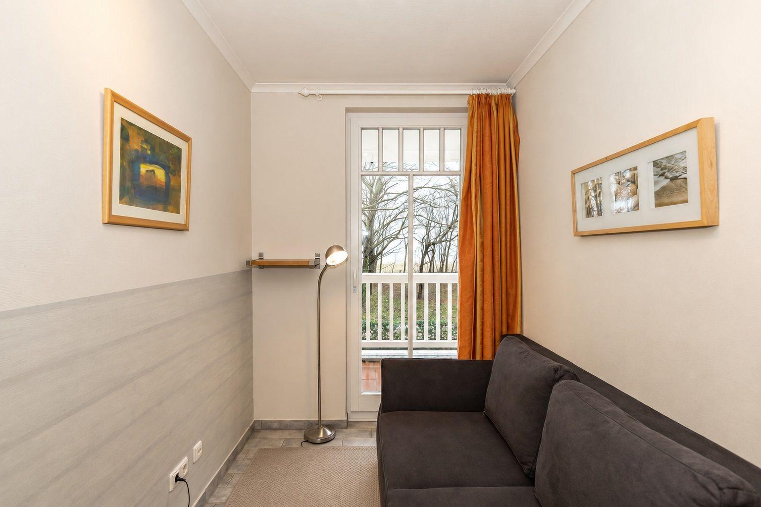 Ein Wohnzimmer mit Sofa, Fenster und Vorhang. Wandbilder und Stehlampe sind sichtbar.