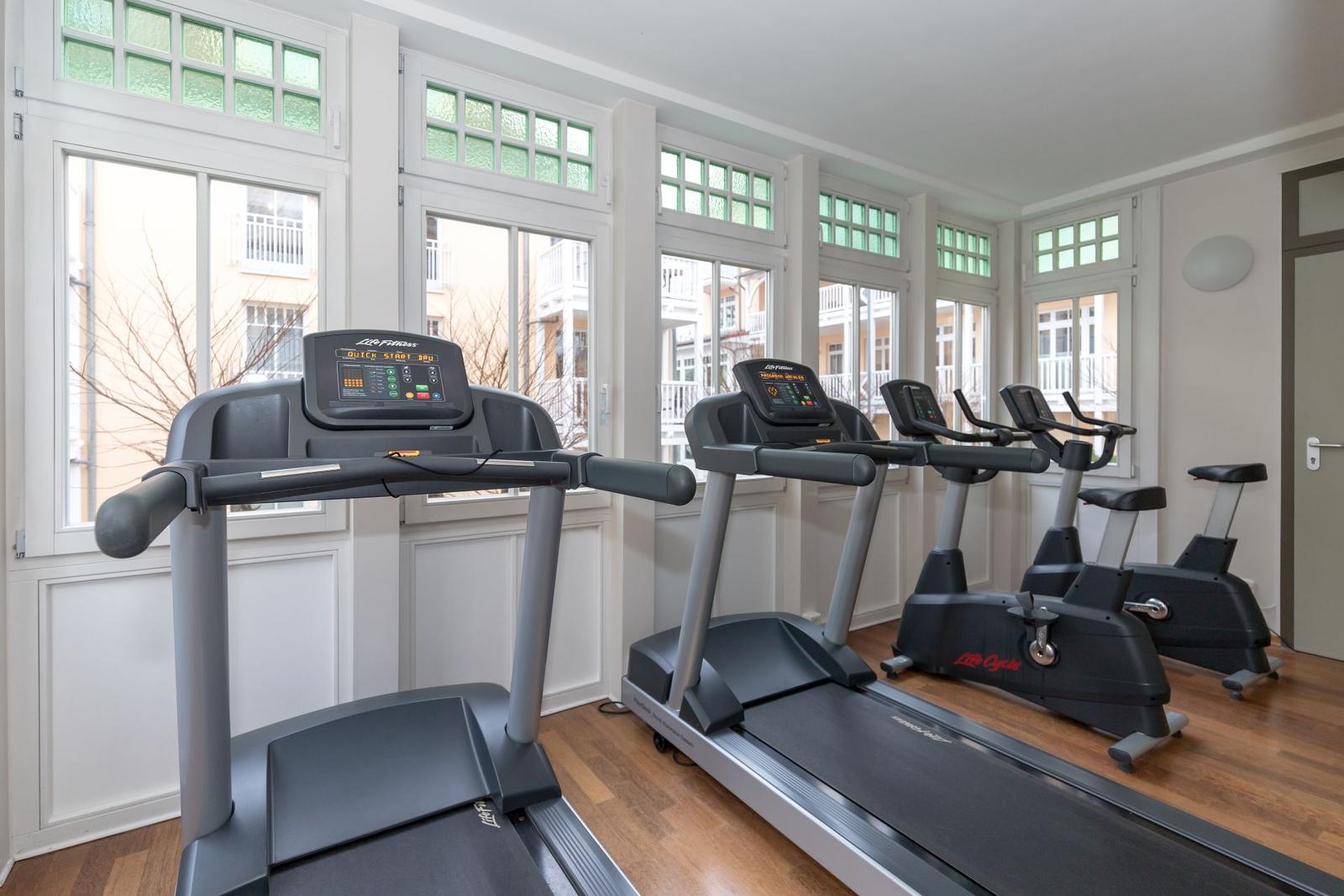 Fitnessraum mit Laufbändern und Fahrrädern vor großen Fenstern.