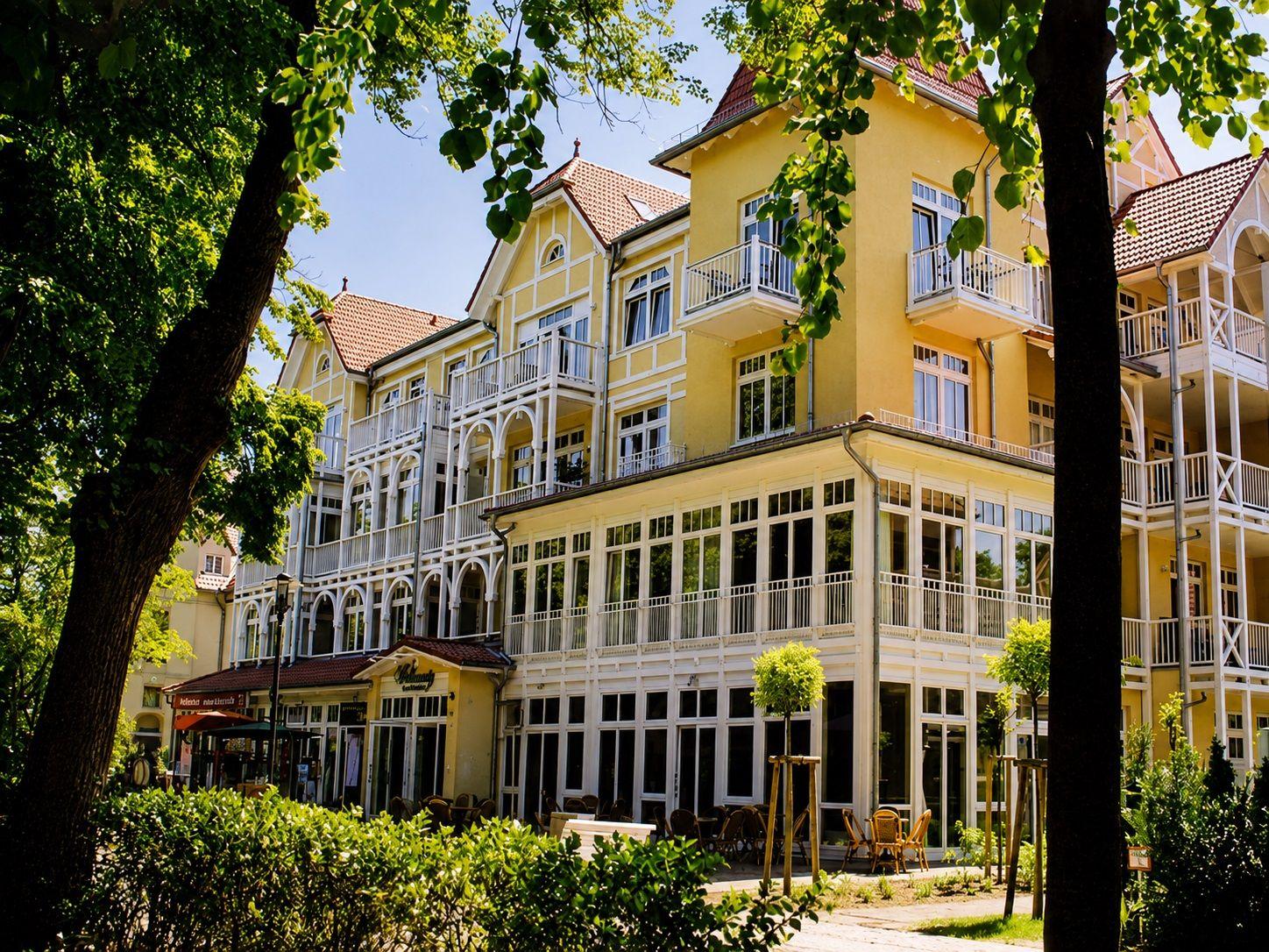 Yellow building with white balconies and red roof tiles surrounded by green trees.