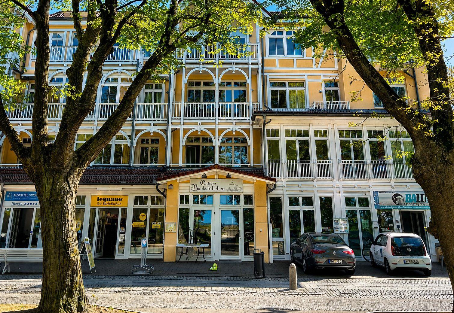 Gelbes Gebäude mit weißen Balkonen und Geschäften im Erdgeschoss unter Bäumen.