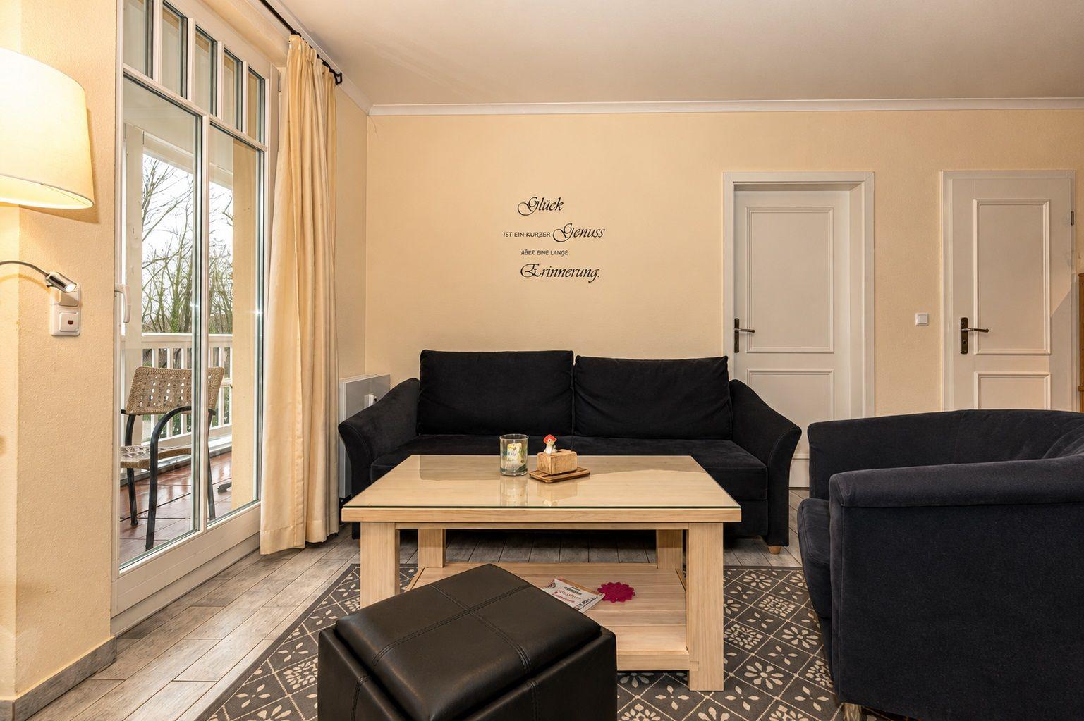 Wohnzimmer mit Sofa, Sessel und Tisch. Durchgangstür und Balkontür. Wanddekoration mit Zitat.