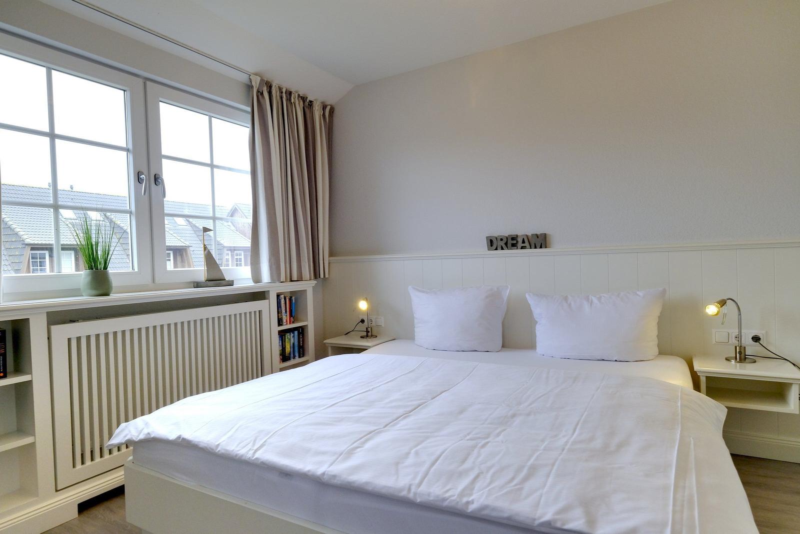 Schlafzimmer mit weißem Bett, Nachttischen und Fenster. Wand mit DREAM-Schriftzug.
