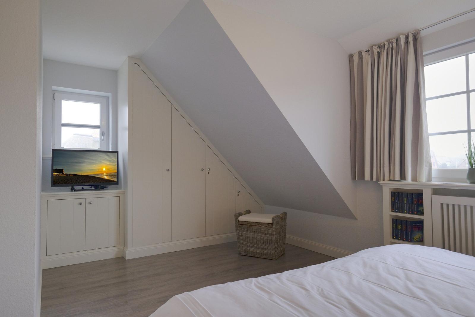 Schlafzimmer mit Dachschräge, Bett, Schrank, TV und Fenster mit Vorhängen.