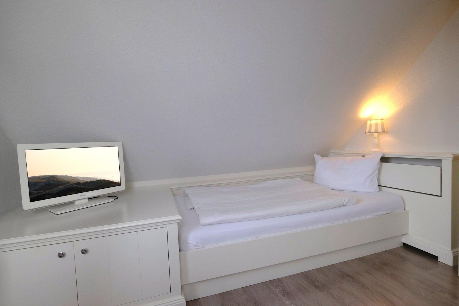 Schlafzimmer mit weißem Bett, Fernseher und Nachttisch unter schräger Decke.