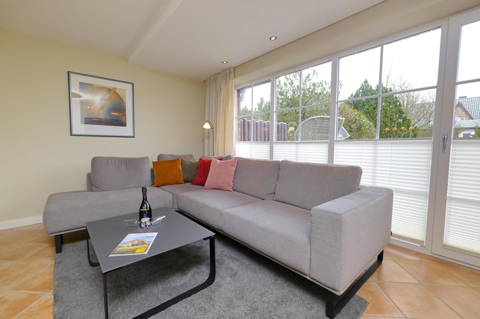 Ein Wohnzimmer mit grauem Sofa, Kissen und Couchtisch. Große Fenster mit Blick auf Garten.