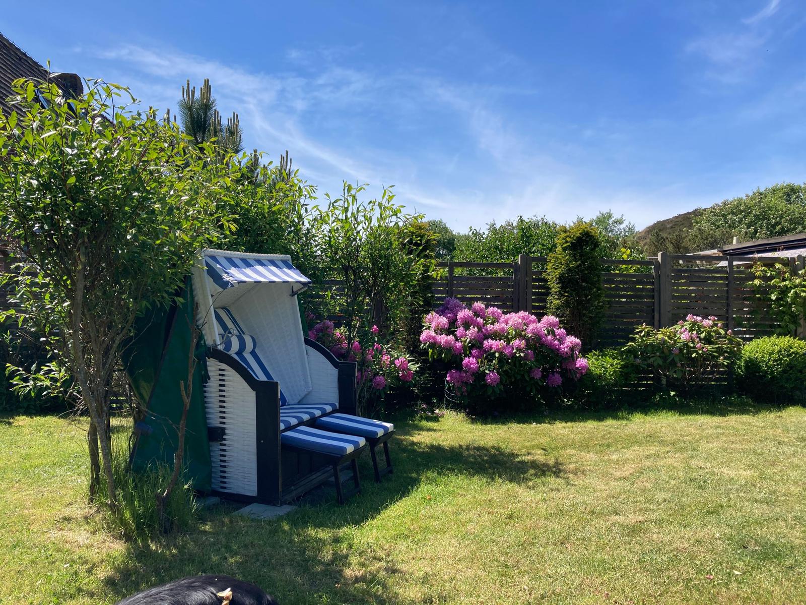 Hinterhof mit Strandkorb, Blumen und Grasfläche unter blauem Himmel.