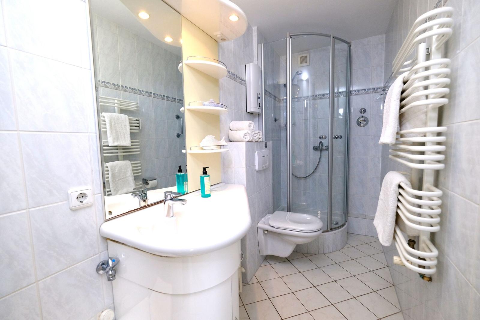 Badezimmer mit Dusche, Waschbecken und Toilette. Weiße Fliesen und Handtuchheizung.