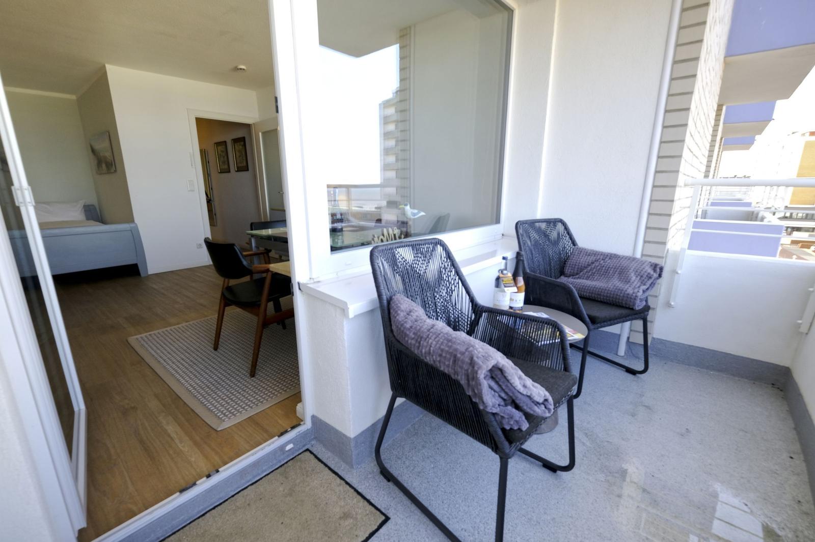 Balcony with two chairs and a table. Glass door leads into a room with wooden floor.