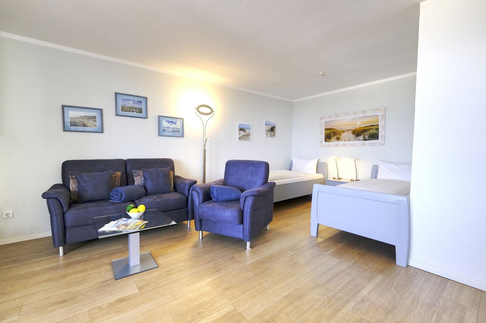 Wohnzimmer mit Sofa, Sessel und zwei Betten. Holzboden und Wandbilder.